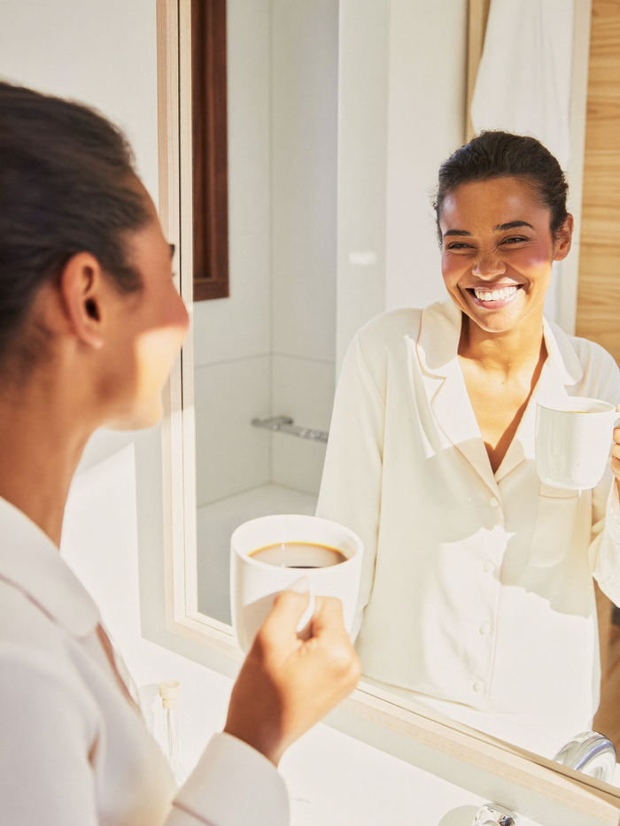 Une femme sourit à son reflet dans le miroir de la salle de bains, une tasse de café à la main